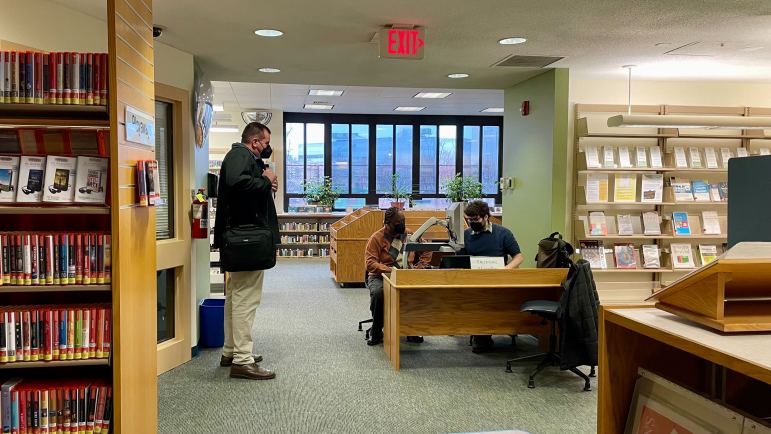 Two people sit at a desk in a library, looking at a laptop screen.