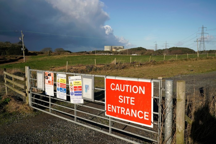 Photo of a gate with a sign saying ‘Caution site entrance’ and a view of Wylfa Nuclear Power Station in the distance 