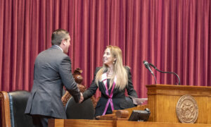 A woman in a black and pink blazer shakes the hand of a man in a suit. There is an ornate podium in front of them and red curtains behind