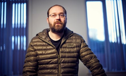 Portrait of Phil Lewis in a brown hooded coat, with an out of focus background of large windows with blue vertical blinds