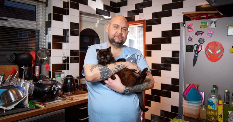 Martin Ives a bald man wearing a blue tshirt, is holding a cat in his kitchen