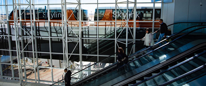 The Skylink transit system that moves people from concourse to concourse at the DFW International Airport is shown outside through windows next to indoor escalators.