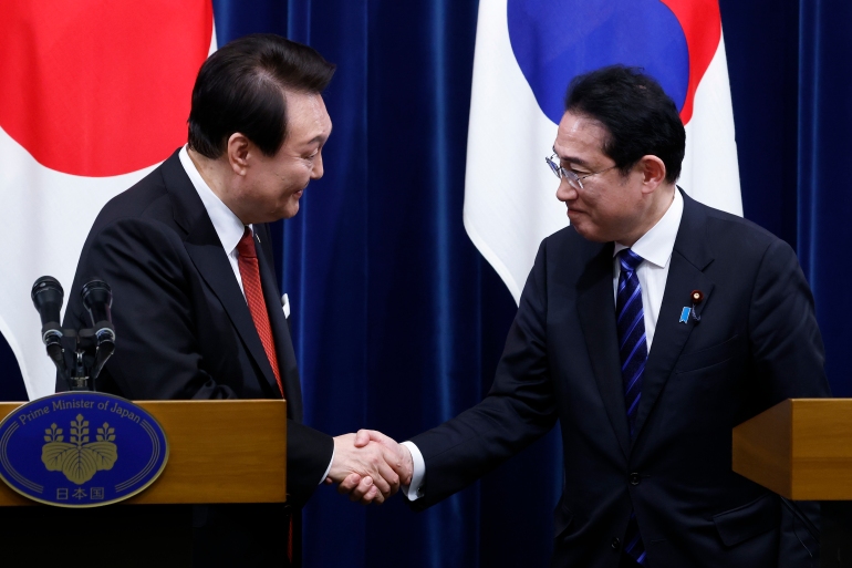 South Korea's Yoon Suk-yeok and Japan's Fumio Kishida shaking hands and smiling in front of the two countries' flags.