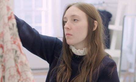 A woman with long hair and a high frilly collared shirt under a jumper looks at a dress