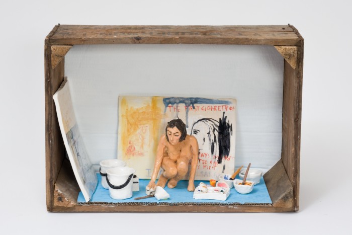 A small naked ceramic woman crouches in her artist’s studio in an open crate