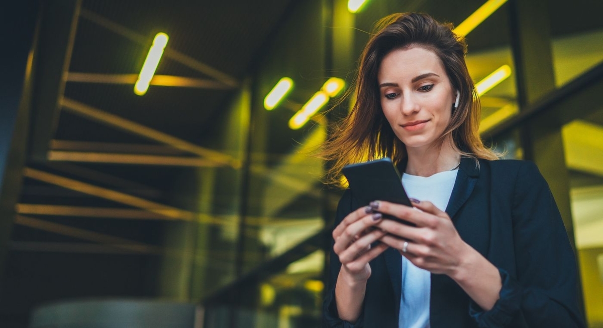 Woman in a black blazer holding a smartphone in front of a building with a lot of lighting.