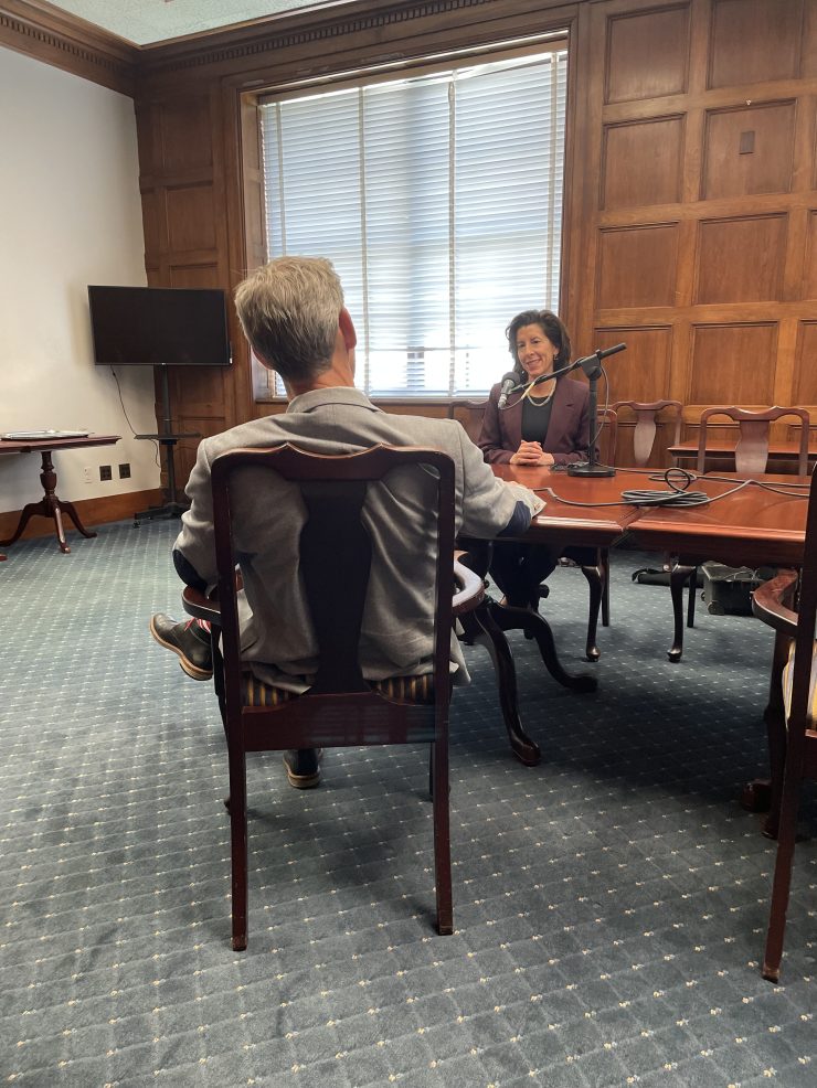 In an official looking room with wood paneling and light blue carpeting, a man, Kai Ryssdal, who has gray hair is seated in a dark wood chair. He is wearing a suit jacket and is facing a a woman, with shoulder-length dark hair, Commerce Secretary Gina Raimondo, who is sitting across a table from him. The two are chatting.