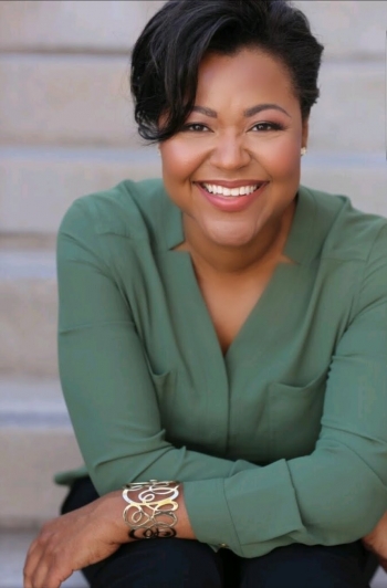 Headshot of a Black woman with short black hair, a green button up blouse and silver bracelet sitting on stairs.