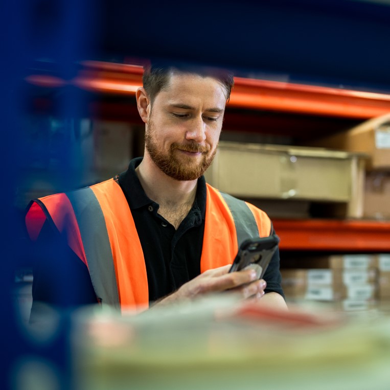 Man wearing an orange vest in a manufacturing or warehouse who appears to be doing inventory or looking at something on the shelf and messaging about it on his phone in Microsoft Teams.