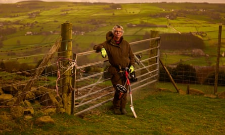 Ruth Harding in a coat, boots and bright green gloves, holding a metal detector and spade, leaning against a fence