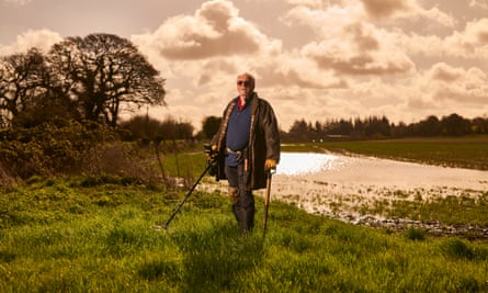 Dave Crisp standing next to a river in the countryside, in a coat, sunglasses and boots and holding a metal detector and a spade