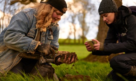 Ellie Bruce and Lucy Gray crouching either side of a hole dug in a field, holding and photographing their find