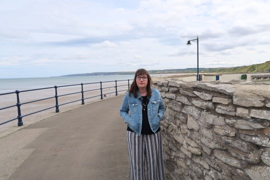 Holly wearing a blue denim jacket and blue and white striped trousers, hands in pockets. She is stood on a promenade with the sea in the background.