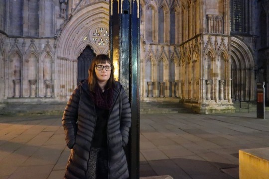 Holly leaning against a lamp post at night with a cathedral in the background. She’s wearing a charcoal coloured coat, burgundy scarf and dark jumper and trousers.