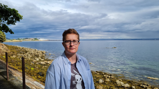 Jacq Kelly outside, by the sea. She's standing on a path. On her left is a rocky, grassy bank (covered mostly with grass, but also assorted grey rocks) which leads down to the sea, which is calm and blue. There's an overcast sky, which makes up most of the top half of the photo; and the edges of a tree overhanging the path to the left of the photo. Jacq is foregrounded in the photo, wearing a blue and white striped shirt over a dark grey t-shirt which has a picture of a small white animal in the centre. Photo is cropped at elbow length. Jacq is wearing glasses and looking directly into the camera.