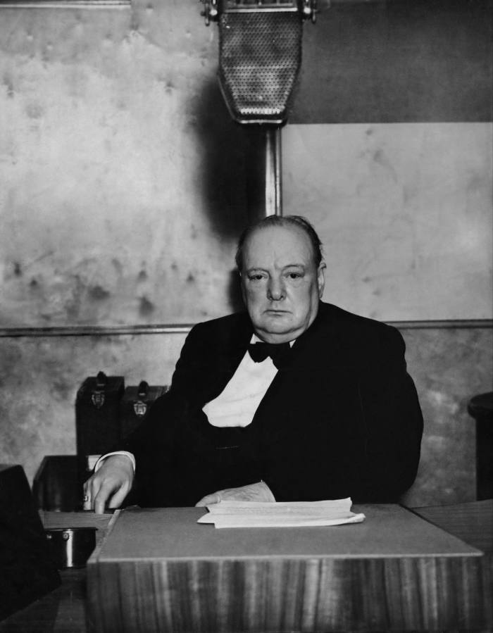 Churchill, in suit and bow tie, sits at table on which there are sheets of paper