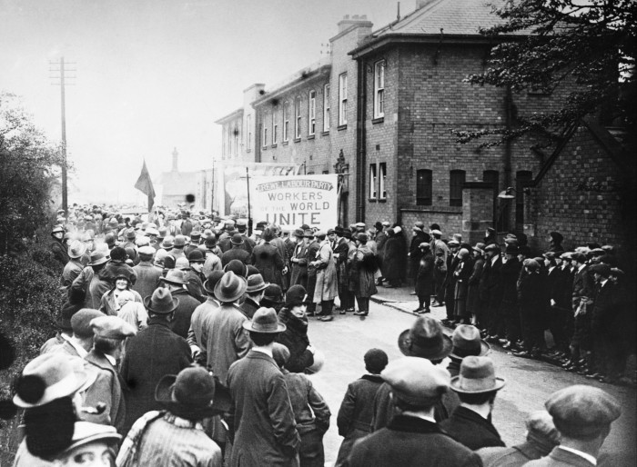 A black and white photograph of a large crowd of people in the street, some of them carrying a placard saying ‘workers of the world unite’