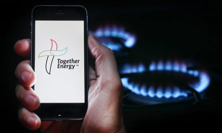 The Together Energy logo displayed on a smartphone being held up to the camera, with lit gas cooker rings behind