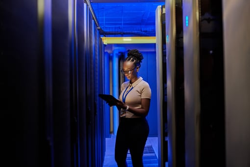 A person standing in a dark server room looking down at a tablet device