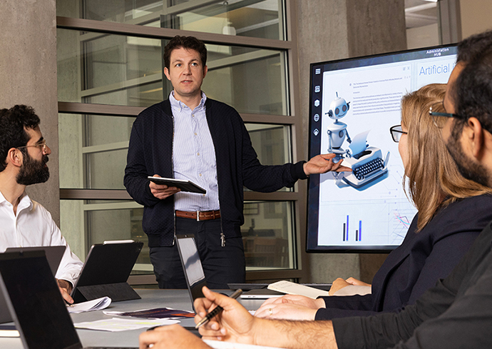 Individual standing at a computer monitor presenting to other individuals sitting at a table.