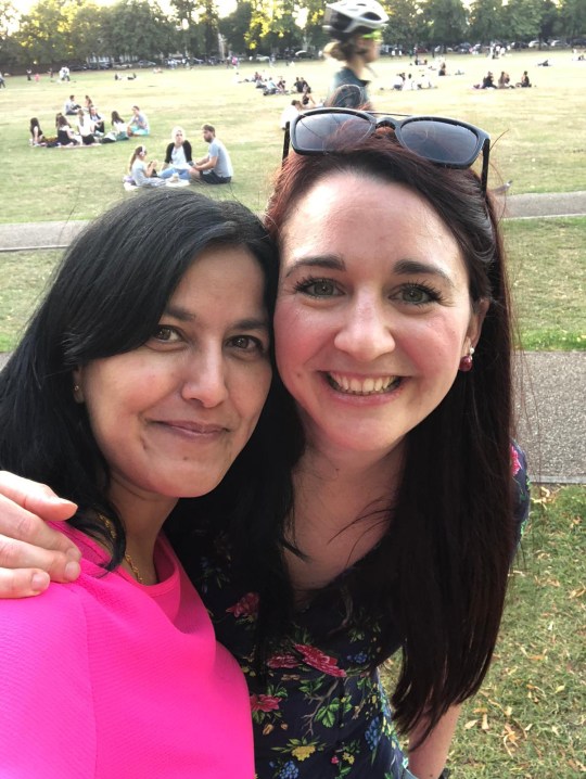 Charlie Bishop in a park with a friend. They are both smiling at the camera, Charlie's arm is around her friend and there's grass/people picnicking behind them.