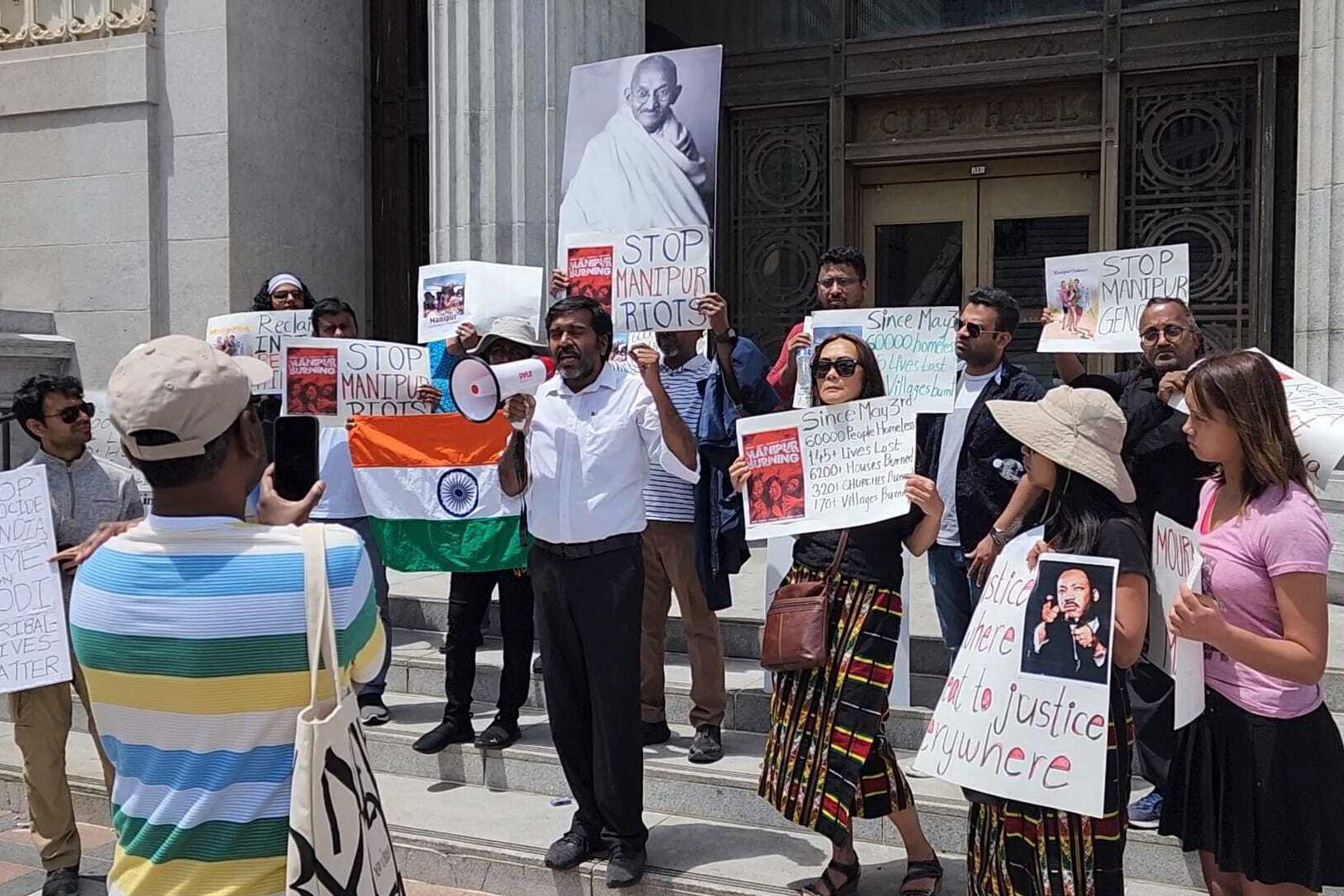 A group of people stands outdoors in front of a building holding signs while one person speaks into a megaphone.