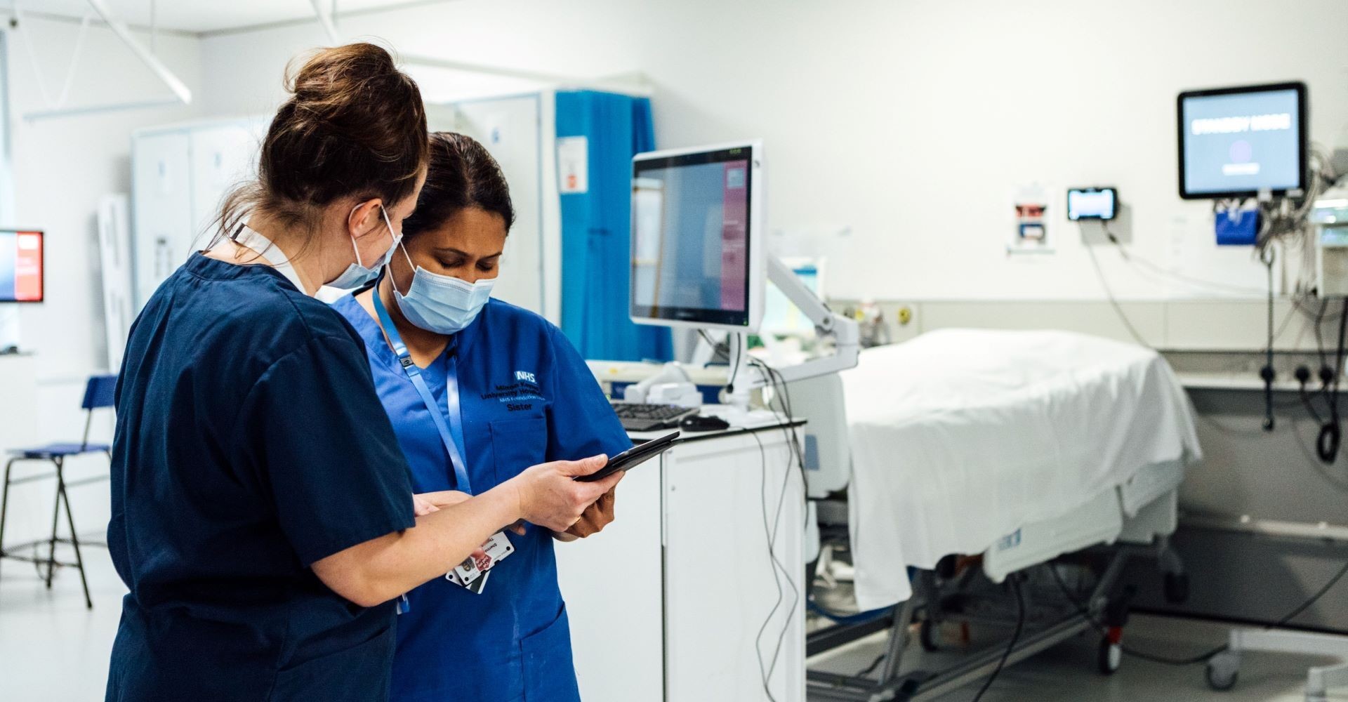 Two medical professionals in a hospital examination room reviewing a patient chart on a mobile tablet device.