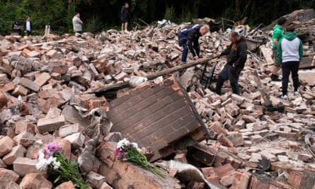 People stand among the ruins of the Crooked House pub, with bunches of flowers in the foreground. 