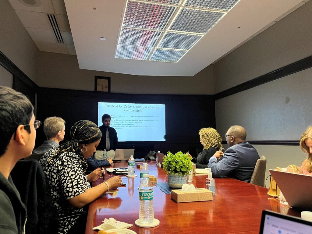 A student stands at a podium in front of a white video screen with a wood conference table of five people in professional attire 