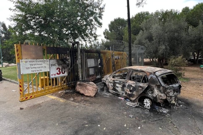 A small car sits charred, from a fire, in front of a yellow gate and and a razor-wire fence.