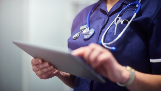 Female surgeon typing on digital tablet in hospital or surgery. She is wearing a dark blue nurse???s top and has her stethoscope around her neck. She is looking at her patients records on her digital tablet and sending emails.