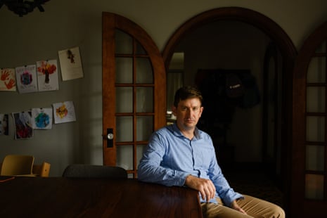 A white man in a blue Oxford shirt and khakis sits on the arm of a couch in a living room, with kids’ art displayed on the wall behind him.