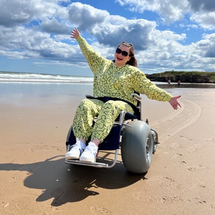 Amy holds out her arms, looking happy, on a beach in the sunshine, in a special chair with huge wheels