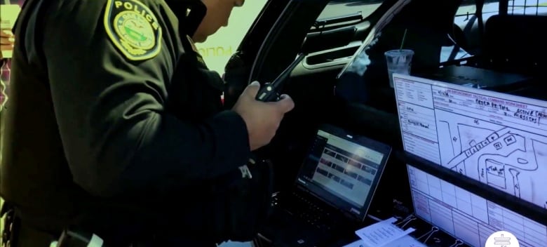 A police officer uses a computer and walkie talkie while looking at a diagram in his trunk.