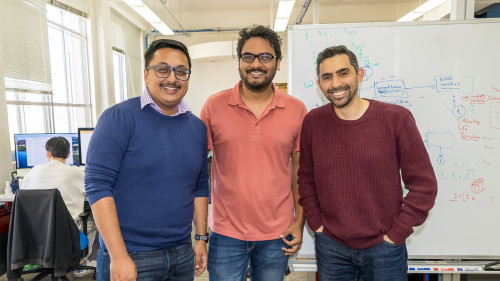 Three men stand next to each other and smile to the camera. They're standing in front of a dry erase board covered in handwritten diagrams. They're in a brightly lit room with others working at computers behind them.