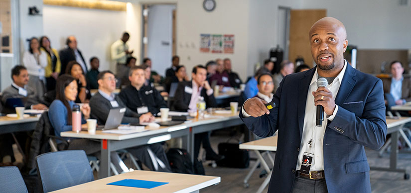 A man speaking at a meeting with seated people in the background.