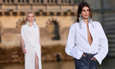 Kaia Gerber wears an unbuttoned white shirt, huge dangling rhinestone earrings like crystal chandeliers, and blue jeans on an outdoor catwalk at Château de Chantilly, France