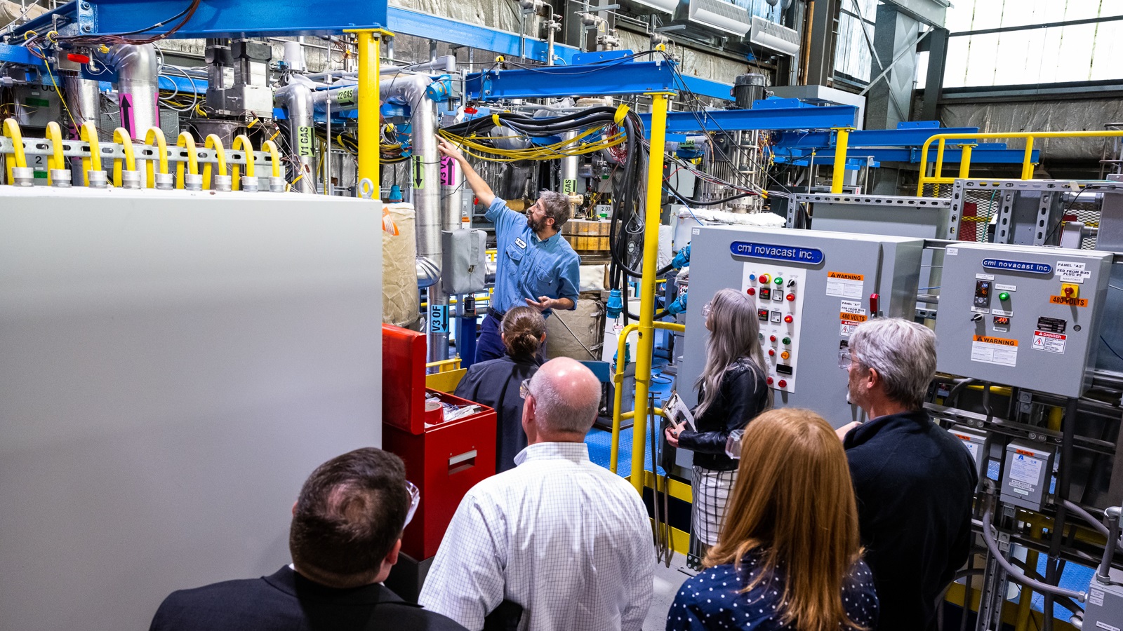 Man in safety glasses points to and explains fast reactor test equipment to small group of visitors.