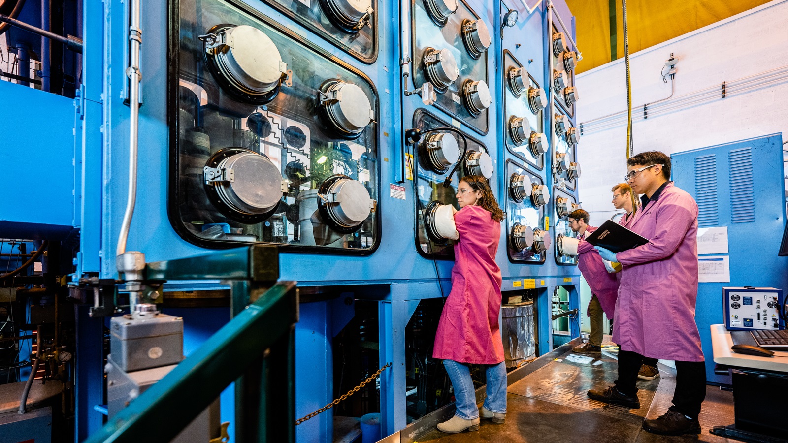 Scientists in magenta coats work in tall glove box laboratory facility.