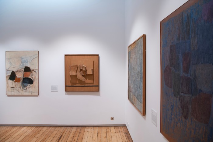 A square relief made of wood with protruding abstract shapes hangs alongside abstract paintings in a gallery