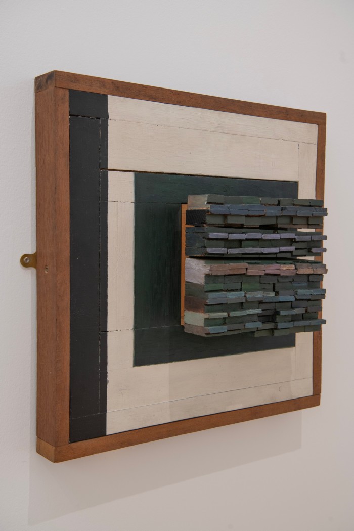 Small pieces of painted wood stuck together form a square shape that protrudes from a larger square slab of wood painted in green white and black 