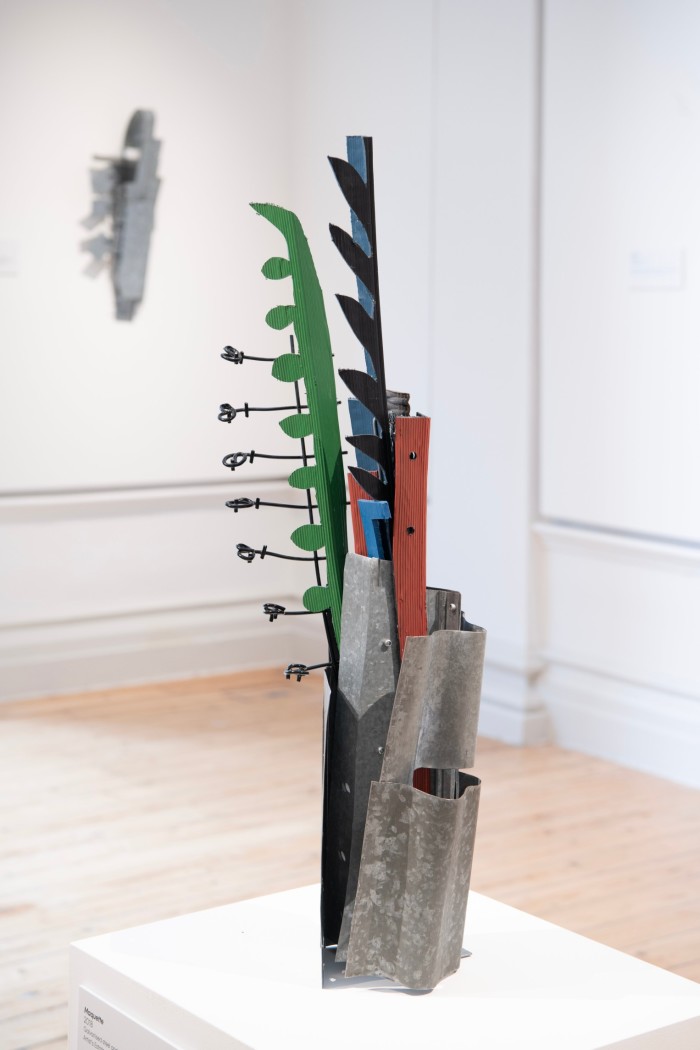 An large abstract metal sculpture on a white plinth with long green, black and red protrusions rising from a metal base