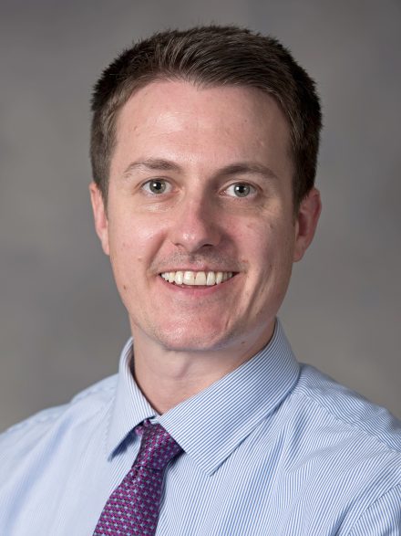 Alex Luchsinger smiles in a headshot against a gray background.