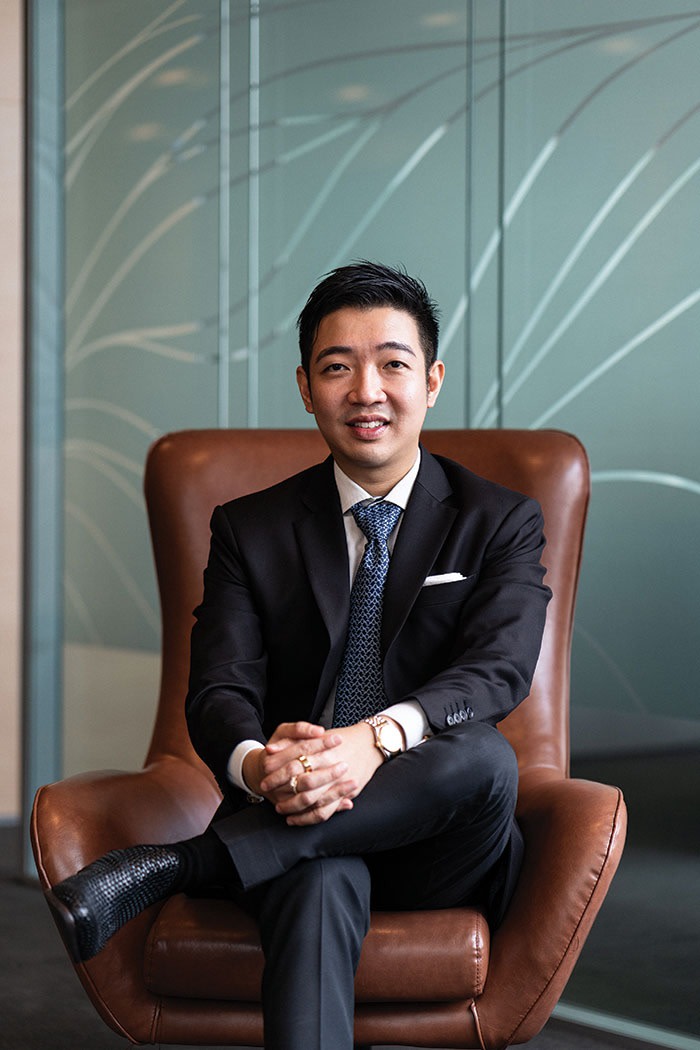 Ivan Cheong, wearing a suit, sits in an office chair, his legs crossed and hands clasped over one leg