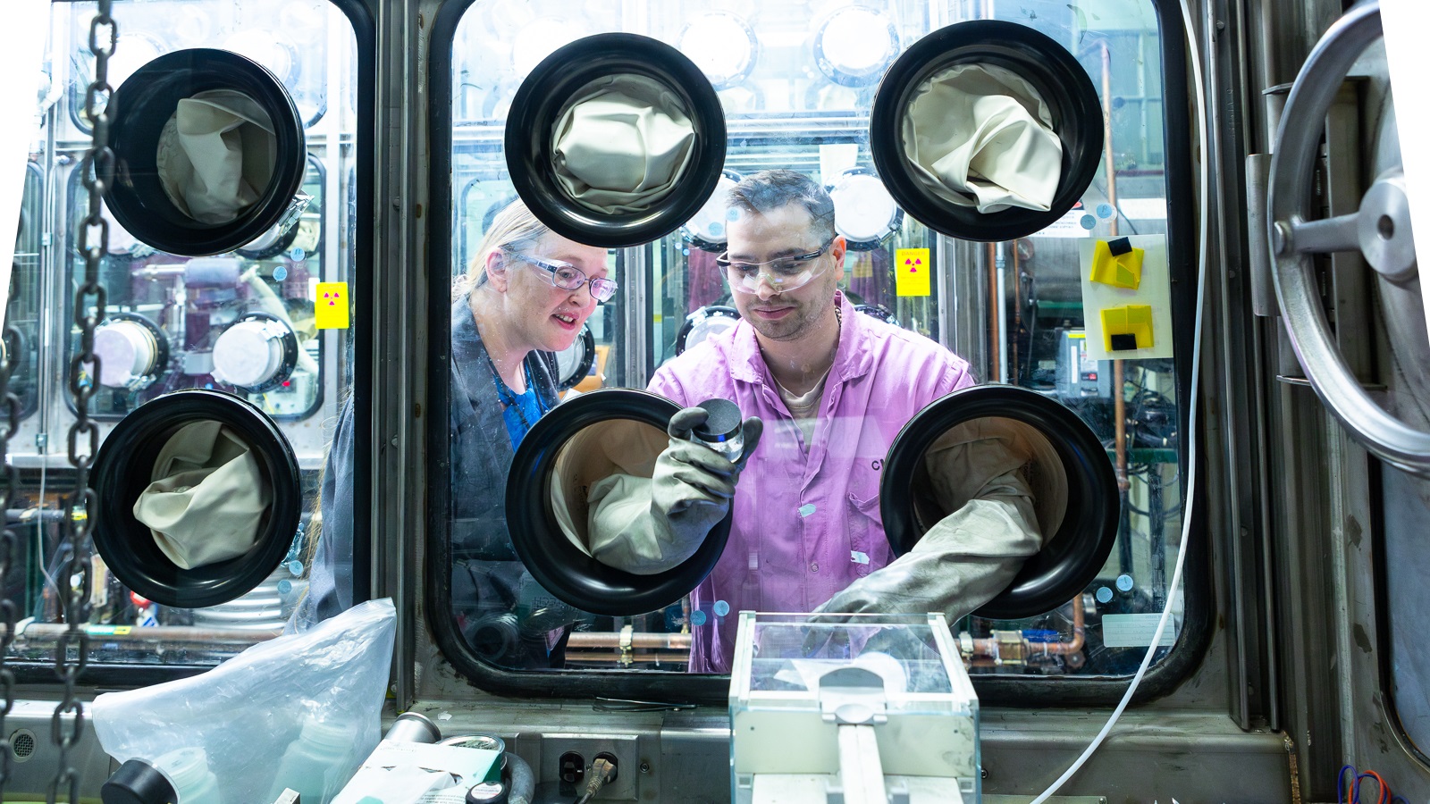 Woman and man in magenta lab coats work with green salt in glass-encased glove box.