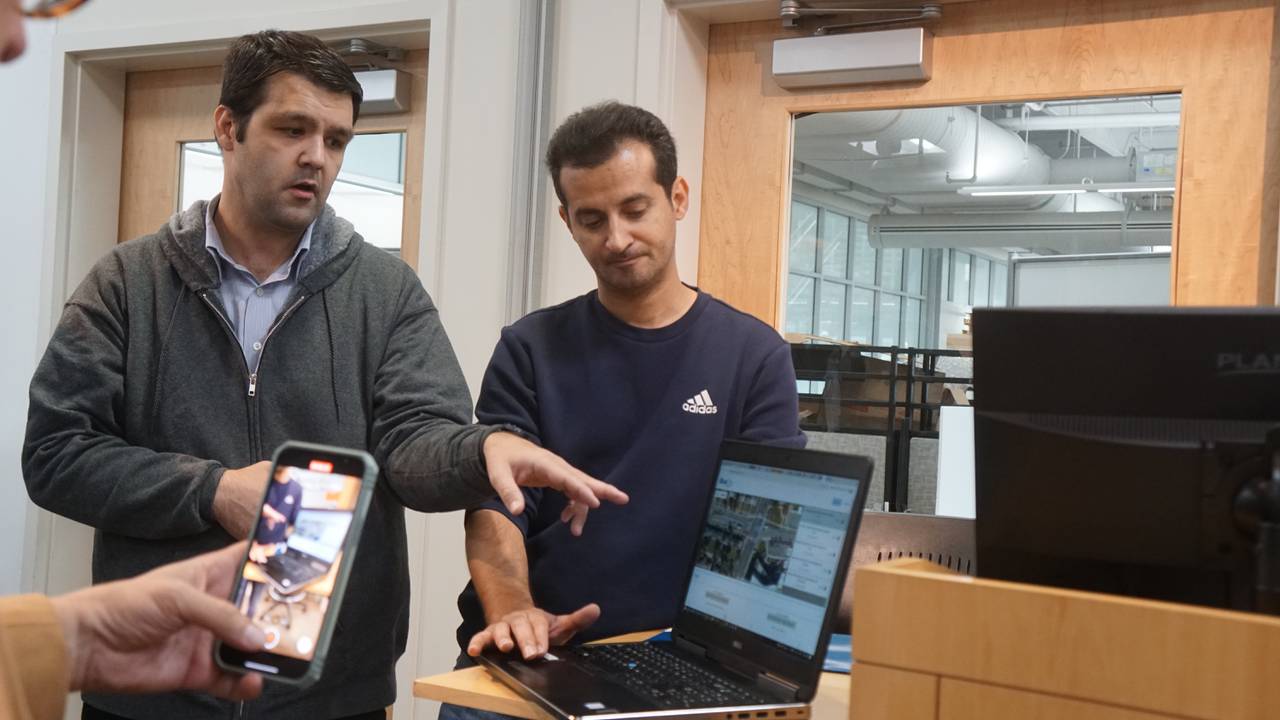 Two men stand in front of a laptop resting on a brown desk and talk to other people standing around them.