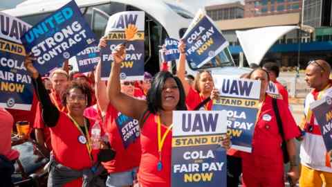 People in red T-shirts hold up placards with ‘UAW’ and ‘cola and fair pay’ written on them at a strike rally