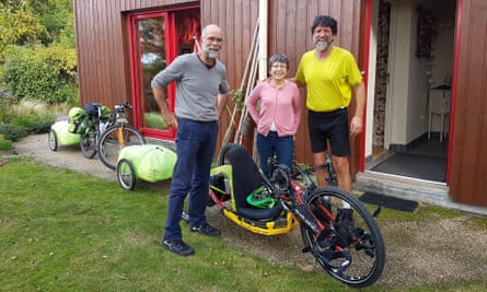 In front of a house, Vincent Berthelot, with a beard and in a T-shirt and cycling shorts next to his recumbent bike, grins as he puts his hand on the shoulder of a woman standing next to a man