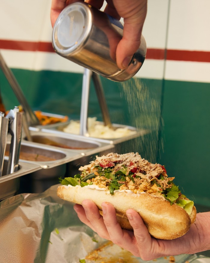 A hand holding Dom’s Subs’ The Grapow sandwich, is filled with spicy Thai ground chicken, gem lettuce, Kewpie mayonnaise, pickled cucumbers and carrots, Thai basil, coriander, pickled red chillies, shallots and toasted rice powder