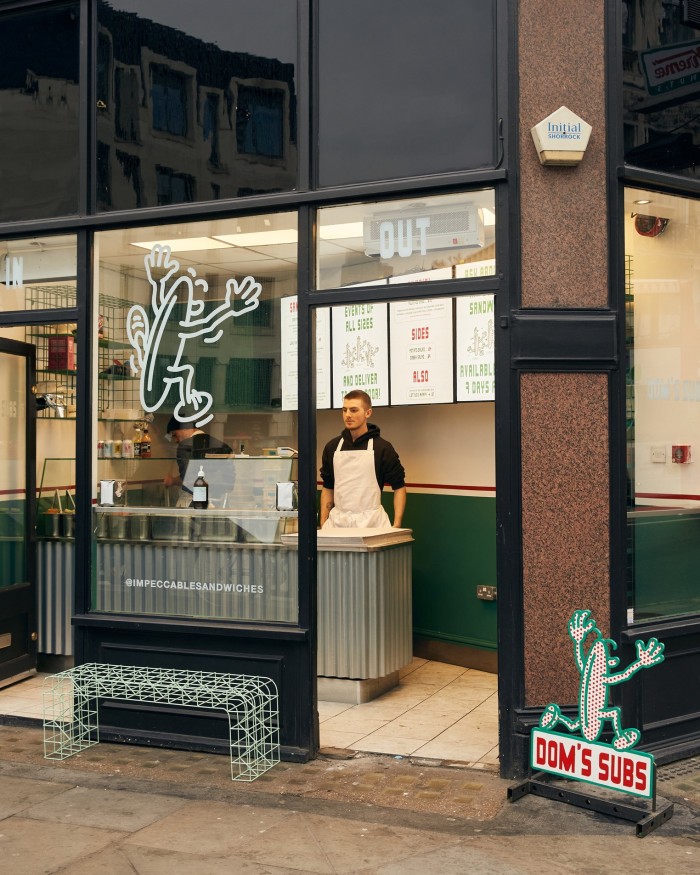 Dom’s Subs Ludgate Circus branch as seen from the outside, with a male employee standing behind a corrugated-iron counter in front of white and dark-green walls